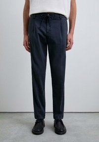 Pantalones navy con cintura ajustable, frente plisado, piernas cónicas y tejido suave, combinados con zapatos negros. Diseño minimalista.
