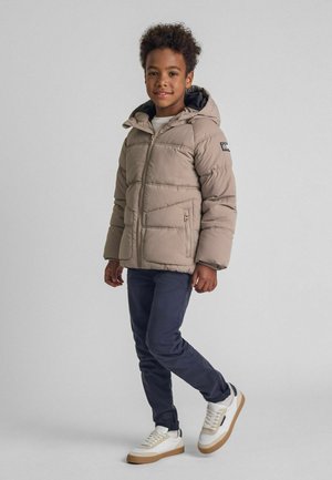 Bežinė pūkinė striukė su gobtuvu ir įstrižomis siūlėmis, derinama su tamsiai mėlynomis kelnėmis ir baltomis sportinėmis bateliais su rudu padu. Lygus audinys ir užtrauktuko detalė.