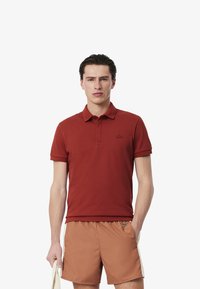 Röd polo-shirt med korta ärmar, krage och broderad logotyp. Kombinerad med ljusbruna shorts, hållna med ena handen.