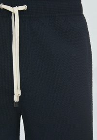 Gros plan sur un pantalon noir texturé avec un cordon blanc noué à la taille et des embouts métalliques argentés aux extrémités.