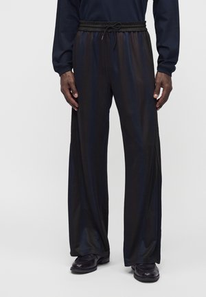 Homme portant un pantalon large en satin noir avec une taille élastique et un cordon, associé à des chaussures noires sur un fond uni.