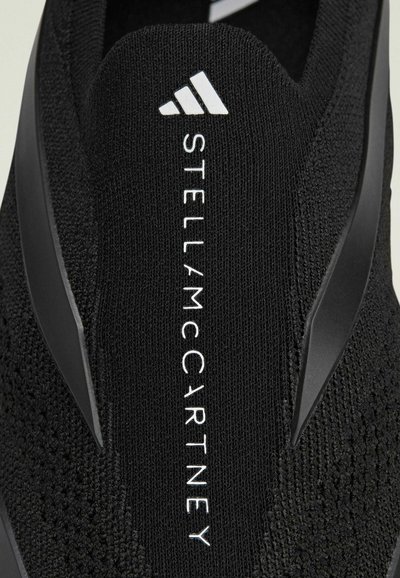 Black knit sneaker featuring a textured upper, sleek design, and a white logo "STELLA McCARTNEY" on the side with plastic accents.
