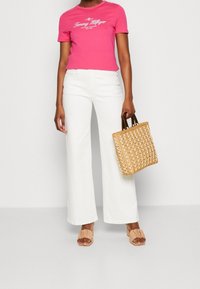 Lyserød cropped t-shirt med hvidt logo, hvide vide bukser, beige flæse-sandaler og en vævet tote taske med mørke håndtag.