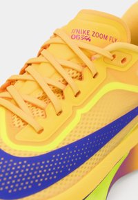 Nike Zoom Fly-sko har en gul syntetisk overdel med blå aksenter, nettingpaneler og en teksturert såle for grep og fleksibilitet.