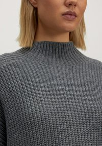 Grauer, strukturierter Strickpullover mit einem hohen, gerippten Kragen. Der Stoff zeigt ein dezentes Muster und wirkt weich und gemütlich.