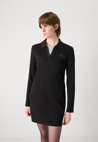 Vestido preto de mangas longas com gola polo, apresentando um tecido texturizado e um detalhe de logotipo no peito. Design simples e ajustado.