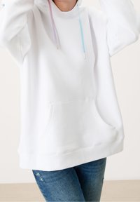 Sweat à capuche blanc avec une poche avant, un ourlet côtelé et des cordons de serrage contrastants en bleu et rose. Texture de tissu lisse. Porté avec un jean.