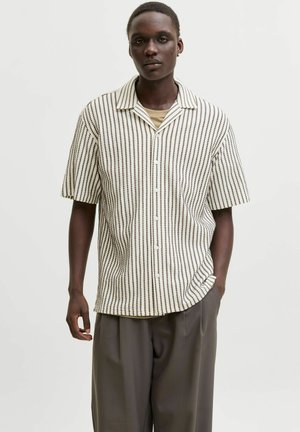 Jeune homme à la peau foncée portant une chemise à manches courtes boutonnée à rayures verticales et un pantalon ample foncé sur un fond uni.