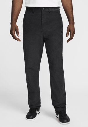 Man die zwarte corduroy broek en zwarte sneakers met witte zolen draagt, staand tegen een effen lichte achtergrond.
