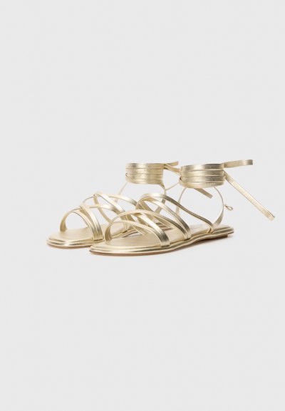 Gullfargede metalliske sandaler med flere slanke stropper og en flat såle. Ankelbånd med knytning for sikker passform. Glatt tekstur.
