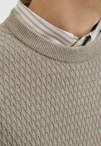 Pull en maille beige clair avec un motif en losanges texturé, doté d'un col rond et superposé à une chemise blanche présentant des rayures grises subtiles.