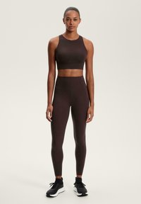Brauner Sportbekleidungs-Set mit einem ärmellosen Cropped-Top und hochgeschnittenen Leggings, aus glattem Material gefertigt, kombiniert mit schwarzen Sportschuhen.