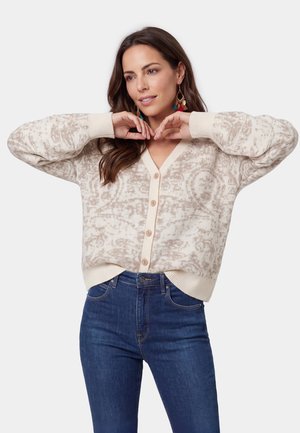 Femme aux longs cheveux bruns portant un cardigan beige à motifs boutonné et un jean bleu, posant avec les mains près du visage.