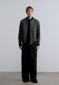 Γκρι μπουφάν τύπου bomber με κλείσιμο με φερμουάρ, συνδυασμένο με μαύρο κοντομάνικο και φαρδιά μαύρη παντελόνια. Μαύρα παπούτσια ολοκληρώνουν την εμφάνιση. Μινιμαλιστικός σχεδιασμός.