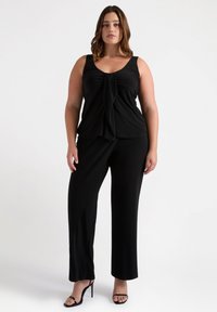 Top negro sin mangas con detalle de nudo drapeado, combinado con pantalones sueltos negros. Tela suave con un ligero brillo y diseño hasta el tobillo.