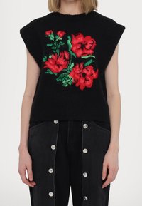Czarny dzianinowy bezrękawnik z czerwonym haftem kwiatowym z zielonymi akcentami, w połączeniu z ciemnymi jeansami z srebrnymi guzikami.