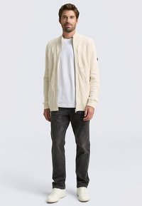 Cremefarbener Cardigan aus Strickstoff mit Diamantmuster, offenem Vorderteil, gerippten Bündchen und Ausschnitt, kombiniert mit einem weißen T-Shirt und dunkler Jeans.