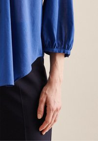 Blusa leggera blu con maniche a sbuffo, orli rifiniti e una silhouette fluida. Abbinata a pantaloni scuri, che mettono in mostra dettagli lavorati a mano.