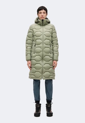 Persona che indossa un lungo cappotto trapuntato verde chiaro con cappuccio, pantaloni blu scuro e scarponi da trekking, in piedi su uno sfondo chiaro uniforme.