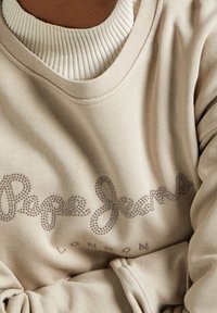 Sudadera beige con un escote en ribete blanco, que presenta un logotipo "Pepe Jeans London" adornado con diamantes en un estilo cursiva. Textura suave.