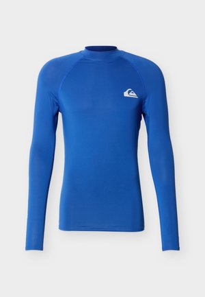 Camiseta deportiva de manga larga de color azul brillante con cuello alto y pequeño logo blanco en el lado izquierdo del pecho sobre fondo liso.