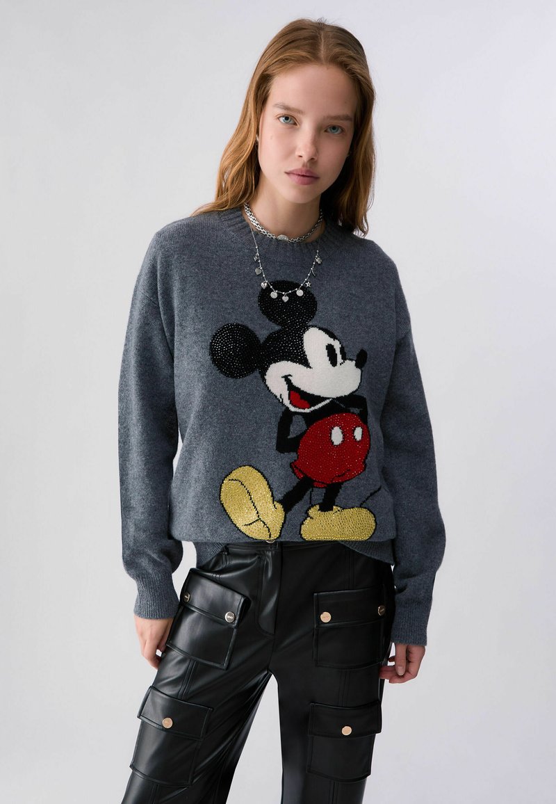 Jeune femme portant un pull gris avec un grand motif Mickey Mouse et un pantalon cargo noir avec plusieurs poches.