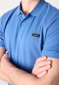 Polo de couleur bleu clair fabriqué en tissu tricoté, doté d'un col, de deux boutons et d'une étiquette de logo Calvin Klein noire sur la poitrine.