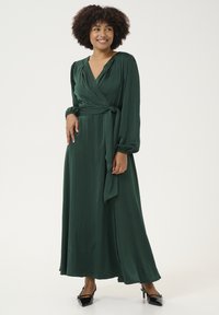 Abito lungo stile avvolgente in satin verde scuro con maniche lunghe e una cintura da annodare in vita. Tessuto liscio; presenta un profondo scollo a V.