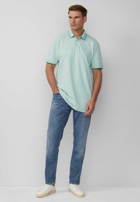 Lichtgroene polo met contrasterende donkergroene kraag en manchetten. G gedragen met een blauwe spijkerbroek en witte sneakers. Eenvoudig ontwerp, klassieke pasvorm.