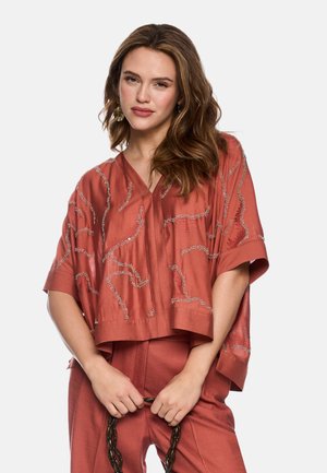 Woman with wavy brown hair wears rust-colored embroidered top and matching pants, holding a black chain-link handle with both hands.