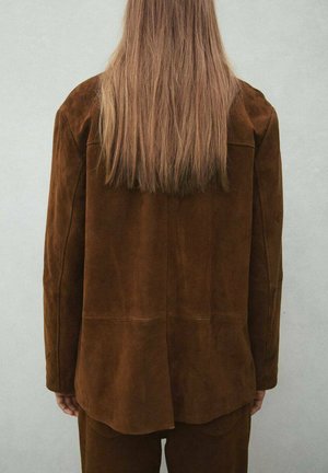 Persona con pelo largo y liso, vestida con una chaqueta de ante marrón y pantalones a juego, de pie frente a una pared lisa de color claro.