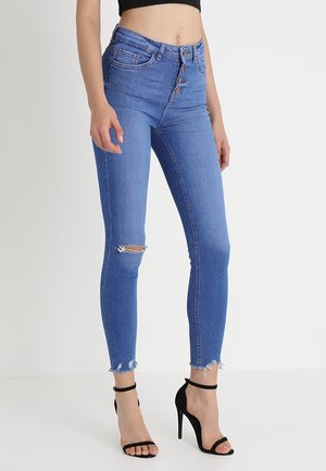 Kobieta ubrana w niebieskie wąskie jeansy z wysokim stanem, z podartym kolanem i postrzępionymi brzegami, zestawione z czarnymi sandałami na wysokim obcasie z paskiem wokół kostki.