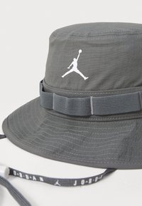 Gray bucket hat made of textured fabric with a wide brim. Features a white Jumpman logo and a dark gray adjustable strap.