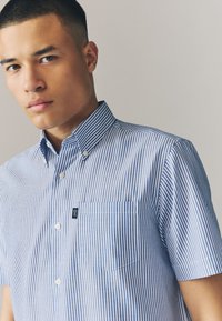 Kortærmet skjorte med knapper, blå og hvide lodrette striber, med en påsyet lomme og et lille logo på brystet. Fremstillet af bomuld.