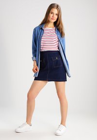 Gonna di jeans con due tasche frontali, di colore blu scuro, abbinata a una camicia a righe rosse e bianche e a una giacca di jeans azzurro chiaro. Sneakers bianche.