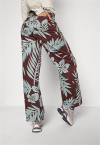 Pantalon à jambes larges en tissu bordeaux orné d'un imprimé de feuilles tropicales et de fleurs bleues. Stylé avec des baskets grises, montrant une coupe décontractée.