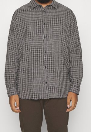 Mężczyzna w koszuli w czarno-białą kratę i ciemnobrązowych spodniach, stojący na białym tle.