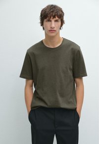 Olive green cotton t-shirt with a crew neckline and short sleeves, paired with dark trousers. Smooth texture, relaxed fit, minimal design details.