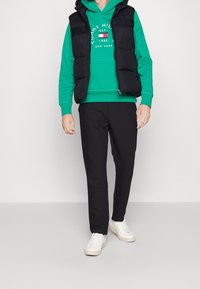 Mężczyzna w zielonym hoodie marki Tommy Hilfiger, czarnej kamizelce, czarnych spodniach i białych sneakersach stoi na tle o jednolitym kolorze.