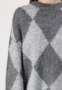 Lähikuva henkilöstä, joka pitää yllään harmaata ja vaaleanharmaata rombikuviollista villapaitaa pyöreällä kaula-aukolla.