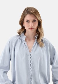 Blusa blu chiaro con bottoni, colletto arricciato, tessuto morbido e bottoni decorativi lungo la parte frontale. Maniche lunghe e ampie.