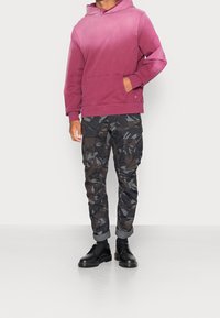 Bordeaux mikina s kapucí a přední kapsou v kombinaci s černými maskáčovými cargo kalhotami s několika kapsami. Černé boty dotvářejí celý outfit.