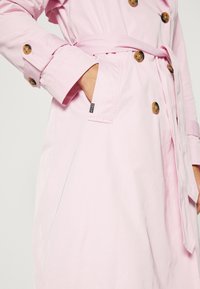 Main dans la poche d'un trench-coat rose clair avec des boutons en écaille et une ceinture nouée à la taille, l'anse de la manche boutonnée au poignet.