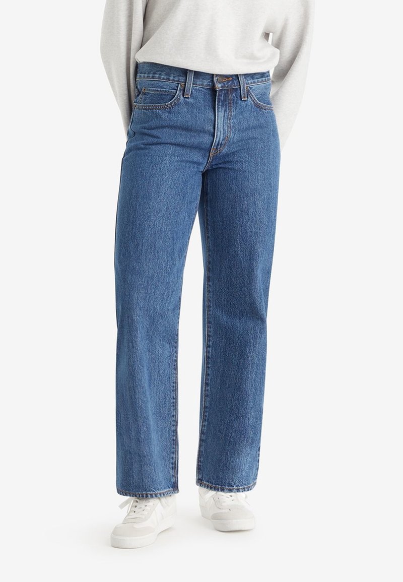 Personne portant un jean bleu à taille haute et jambes droites, un haut à manches longues de couleur claire et des baskets blanches sur un fond uni.