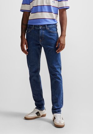 Herren Jeans aus blauen Denim mit geradem Schnitt, klassischem Fünf-Taschen-Stil und dezenten Ausbleichungen; kombiniert mit einem gestreiften kurzärmligen Hemd.