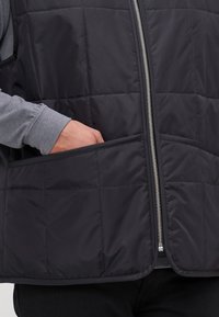Melna aizmugureiztā veste ar kvadrātisku struktūru, priekšējais rāvējslēdzis un sānu kabatas. Valkāta virs pelēka garām piedurknēm krekliņa. Vienkāršs dizains, utilitārs stils.