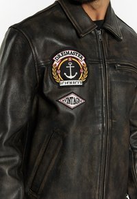 Ruskea nahkatakki, jossa vetoketjullinen etuosa ja suuri kirjailtu ankkurikuvio rinnassa, sekä vintage-tyylisiä yksityiskohtia. Sileä pinta.