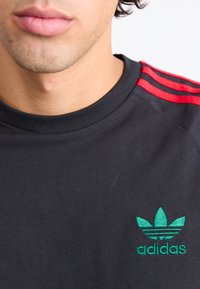 Juodas medvilninis marškinėliai su raudonomis pečių juostelėmis ir žaliu išsiuvinėtu Adidas logotipu su trimis lapų motyvais krūtinės srityje.