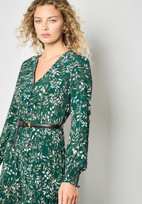 Groene bloemenjurk met lange mouwen, een v-hals en geplooide details. Bevat een dunne zwarte riem en een zachte, gestructureerde stof.