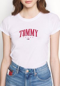 White cotton t-shirt with red "TOMMY JEANS" logo. Short sleeves, round neckline. Worn with light blue denim jeans featuring striped accent pocket.